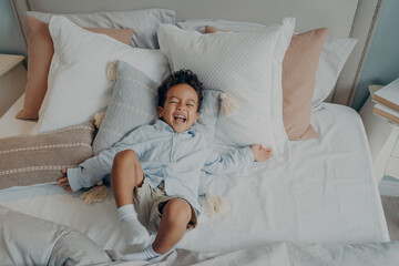 Portrait of afro american joyful little boy on pillows in bed laughing and smiling