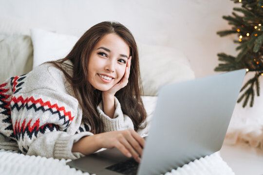 Young asian woman with dark long hair in cozy sweater using laptop on bed in room with Christmas tree at home