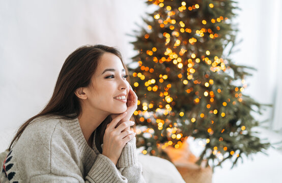 Portrait of young smiling asian woman with dark long hair in cozy sweater in room with Christmas tree