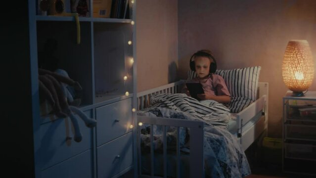 Cute little girl with headphones and tablet listening to music and singing in bed at night. Child is listening to music and playing games or watching video on a tablet