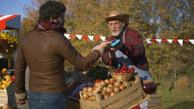 Adult man contactless pays for purchases to farmer using smartphone. Weekend shopping at local farmers market outdoors. Autumn fair. Vegetarian and organic food. Agriculture. Point of sale system.