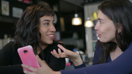 Two female friends talking while looking at smartphone device. Girlfriends chatting and gossiping using phone