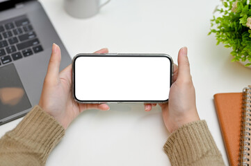 A smartphone white screen mockup in horizontal position is in a woman's hands over office desk