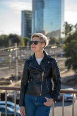 30s woman with short blonde hair in leather jacket in the city with skyscraper on background. Barcelona, Spain