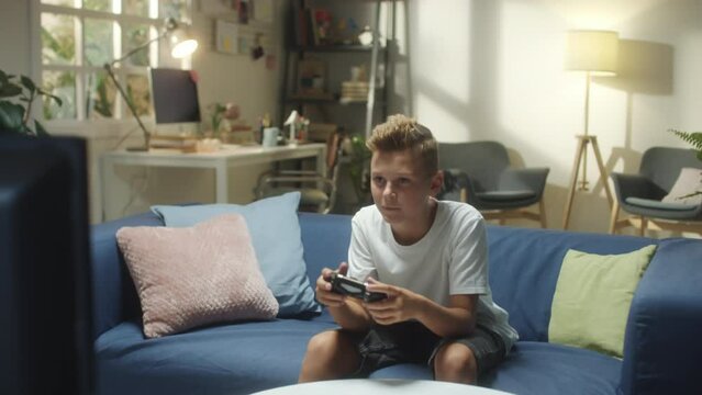 Medium long shot of teenage boy sitting on couch in living room and playing video game on TV with excitement