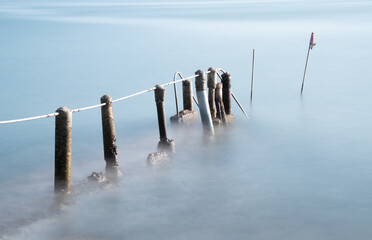 Old broken pier leads into the sea from a beach.  The posts and rails are bent and twisted. Long exposure gives smooth sea surface.
