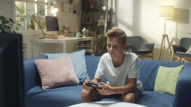 Excited boy sitting on couch in living room and playing video game on TV while spending time at home