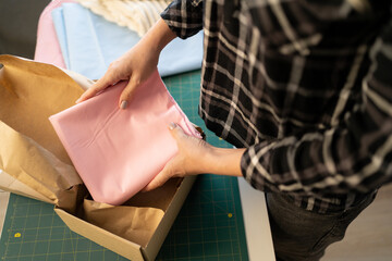 Hands of young female tailor seller of online shop putting folded cloth into box before packing it and sending to client. Small online business owner preparing deliveries in office