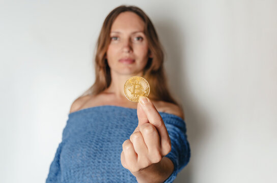 Joyful woman standing holding bitcoin paying attention to new digital cryptocurrency wearing blue sweater. Indoor studio shot isolated on white background.