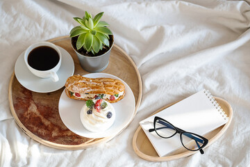 Cakes, coffee cup, notebook and eyeglass on wooden tray on bedding. Cozy breakfast in bed.