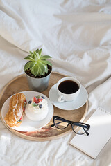 Cakes, coffee cup, notebook and eyeglass on wooden tray on bedding. Cozy breakfast in bed.