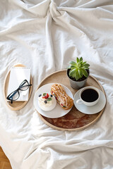 Cakes, coffee cup, notebook and eyeglass on wooden tray on bedding. Cozy breakfast in bed.