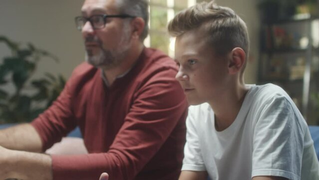 Tilt up shot of mid-aged father and teenage son playing console game together at home