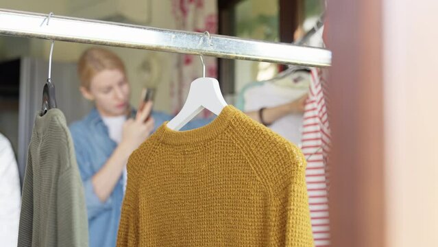 Knitted sweater hanging on rail, woman on background out of focus taking picture of clothes for resale.