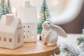 Beautiful holiday card. The concept of a happy new year and Christmas 2023. Christmas trees, a white rabbit and Scandinavian white houses on a wooden table in a cozy living room.