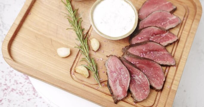  Sliced Beef Meat. Rare medium cooked beef with garlic and rosemary, white sauce and salt in a Woden plate. The salt falls on the meat. Restaurant Food. Rotating Plate on white background