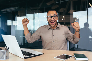 Happy and successful businessman celebrating achievement, portrait of man at work in office looking at camera and holding hands up, celebrating victory and triumph, employee using laptop.