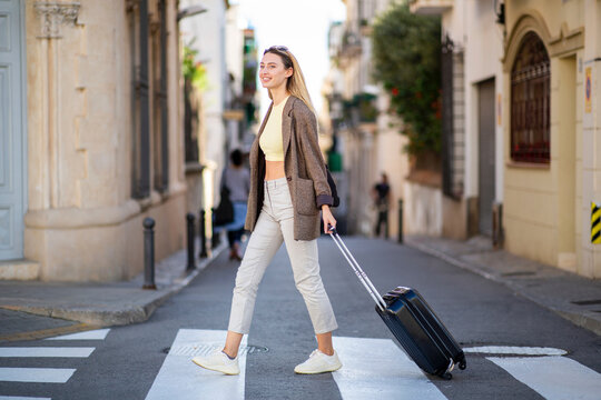 Stylish woman pulling luggage while crossing city street
