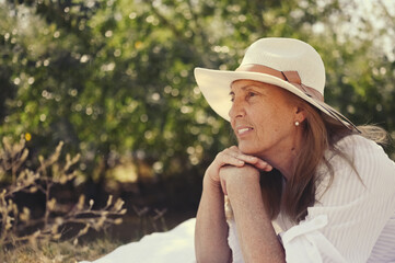 Close up portrait of senior happy woman at summer outdoors. Old elegant lady in straw hat and white shirt lying on grass during picnic at countryside. Active retired people concept