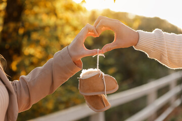 Man and pregnant wife are doing heart gesture with hands and holding warm baby shoes on nature...