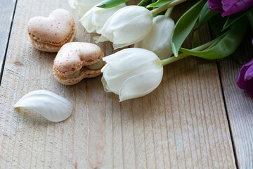 Two tasty creamy macarons are lying on the wooden table with white tulips. Two macarons in form of the heart as 8. Dessert is on the light wooden table