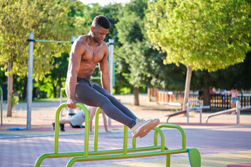 Young fit shirtless black man doing calisthenics workout on parallel bars outdoors on sunny day....