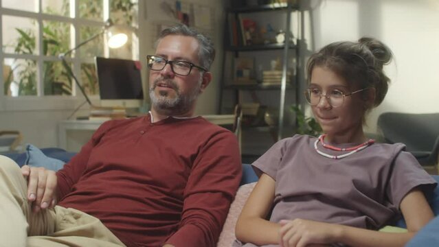 Father And Teenage Daughter Sitting On Sofa In Living Room, Smiling And Chatting While Watching TV Together