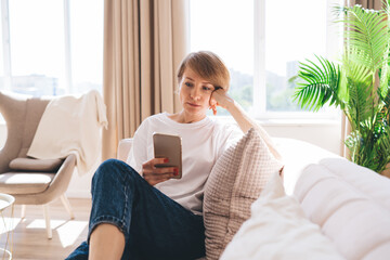Pensive middle aged woman with smartphone relaxing on sofa