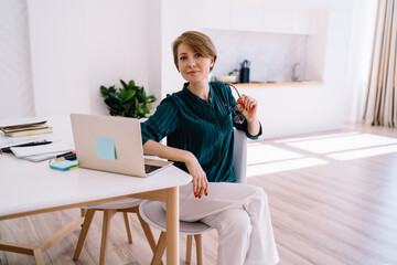 Cheerful adult female freelancer sitting at home with laptop