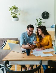 Smiling boyfriend showing calculator to his cheerful girlfriend while doing home finances together online on a laptop computer in the living room