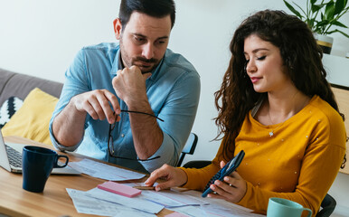 Serious girlfriend showing calculator to her boyfriend while doing home finances together online on a laptop computer in the living room
