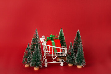 Miniature shopping cart with Christmas trees and gift box on red background with copy space....