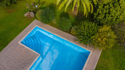 Aerial view of a rectangular swimming pool belonging to a large villa. The pool is empty and you can see the steps. Around the water there is a stone floor and the tropical garden.