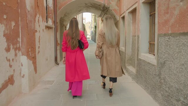 Young women friends in bright colorful coats and fashionable purses walk in shade along narrow Venetian street with old buildings backside view