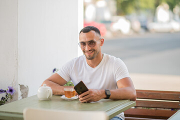 a happy man in a white T-shirt, talking on a cell phone, sitting at a table in a cafe on the street.A man has breakfast and drinks tea in a cafe, Freelance office business