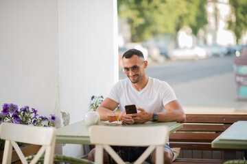 a happy man in a white T-shirt, talking on a cell phone, sitting at a table in a cafe on the street.A man has breakfast and drinks tea in a cafe, Freelance office business