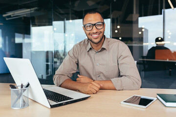 Fototapeta na wymiar Portrait of successful investor, african american man smiling and looking at camera, man working inside office using laptop at work, satisfied with achievement result.