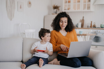 Cheerful young Italian woman with curly hair dressed in orange blouse and blue pants sitting on sofa using laptop, working at home at the kitchen with son playing on phone, talking with mom. Family