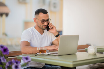 A man is sitting in a city cafe outdoors with a phone and laptop and a cup of tea. A remote workplace for a freelancer. A young guy in a white T-shirt and glasses is talking on the phone