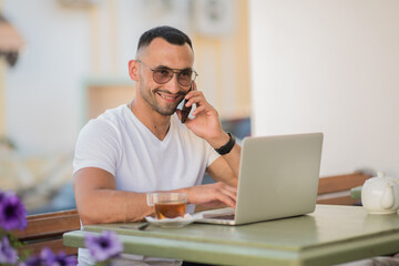 A man is sitting in a city cafe outdoors with a phone and laptop and a cup of tea. A remote workplace for a freelancer. A young guy in a white T-shirt and glasses is talking on the phone