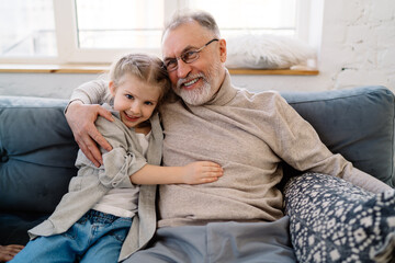 Smiling grandfather and granddaughter cuddling on sofa at home
