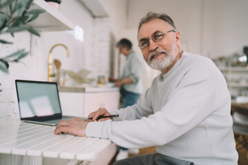 Obraz premium Senior man sitting at table and working on laptop