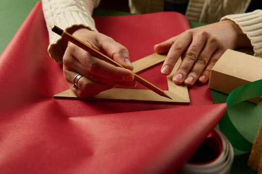 Woman's hands with wooden crayon and triangular ruler draw on wrapping paper, calculate amount of it for wrapping gifts