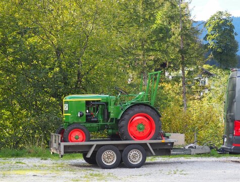 Old green tractor with red wheels on a car trailer with lush trees in the background