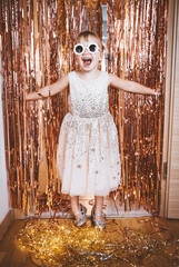 Happy little stylish girl in shiny dress having fun. Festive background with foil curtain...