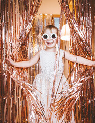 Happy little stylish girl in shiny dress having fun. Festive background with foil curtain decorations for kids birthday or fancy dress party, disco music or New Year.