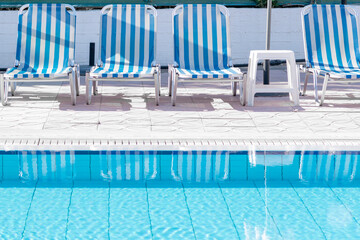 Retro blue and white sunbeds on the poolside of a swimming pool filled with clear blue water at the...