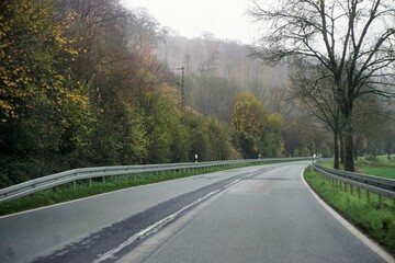 Nasse kurvige Straße  zwischen Ackerfeld und Wald vor weißem diesigen Himmel bei Regen und Nebel...