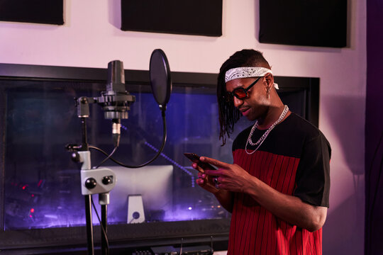 Black male singer browsing smartphone while standing near microphone in studio