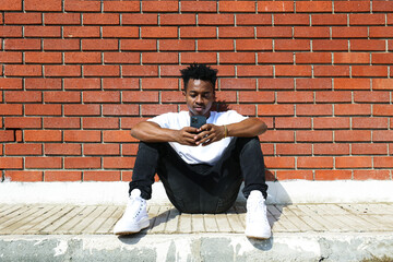Young black man on the street sitting on the curb with his phone and texting. Portrait of a guy wearing a casual outfit leaning on the red brick wall with copy space for text. Background.